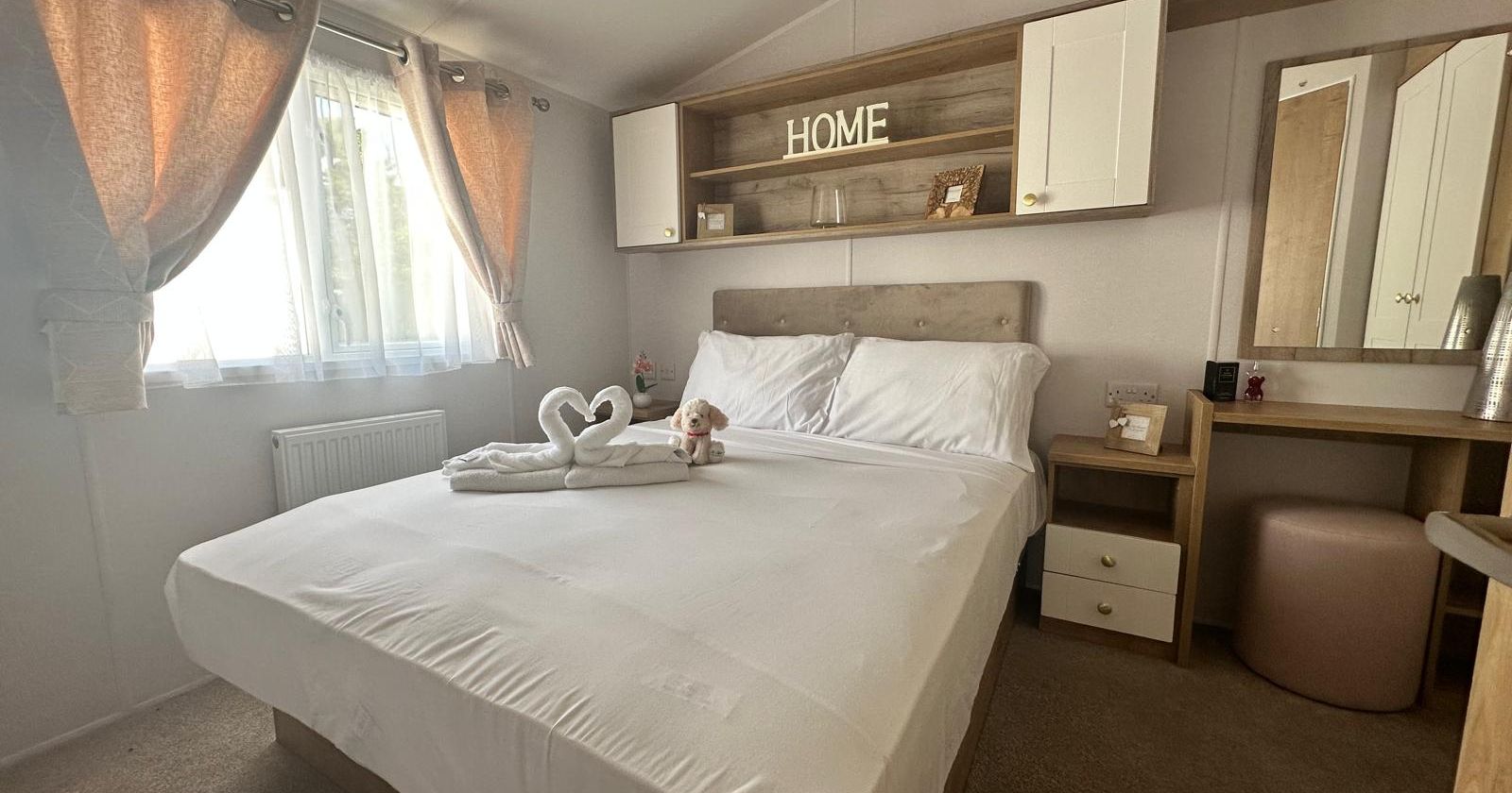 Bright double bedroom with made bed, towel swans, window with curtains, and built-in shelving above the headboard