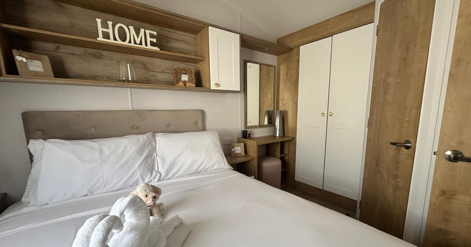 Neatly made double bed with white bedding and a towel swan in a modern bedroom with wooden shelving, vanity mirror and white wardrobe.