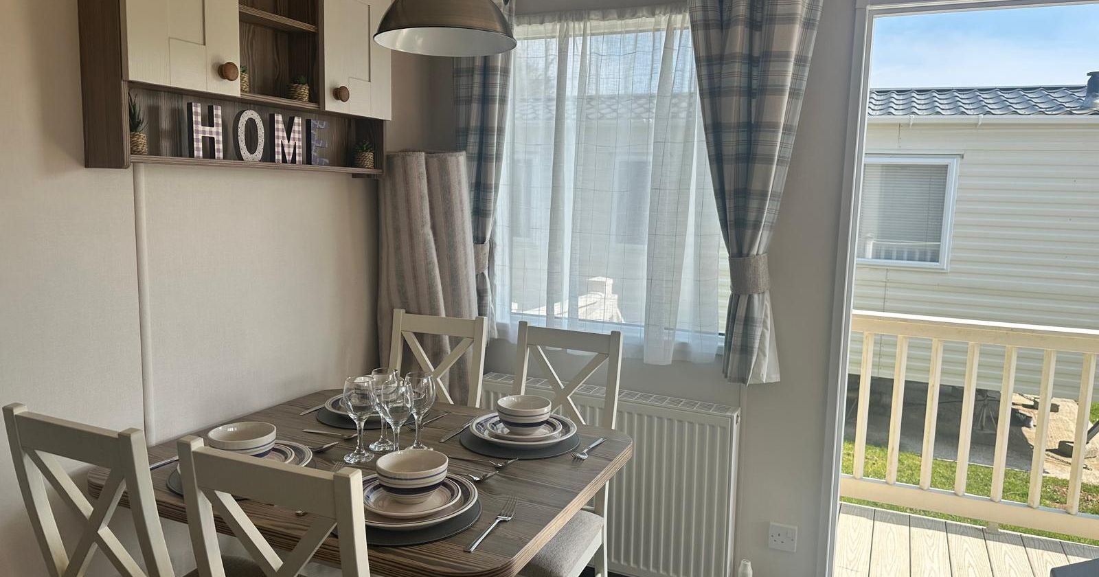 Dining area with a table set for four, grey pendant light, wall shelving and a window with checked curtains beside a glass door to a deck
