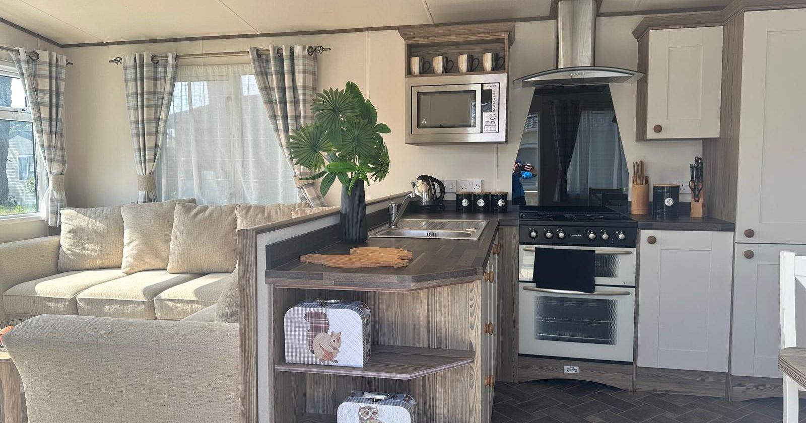 Cosy static caravan kitchen and lounge with grey units, cooker, microwave, and beige sofa by curtained windows