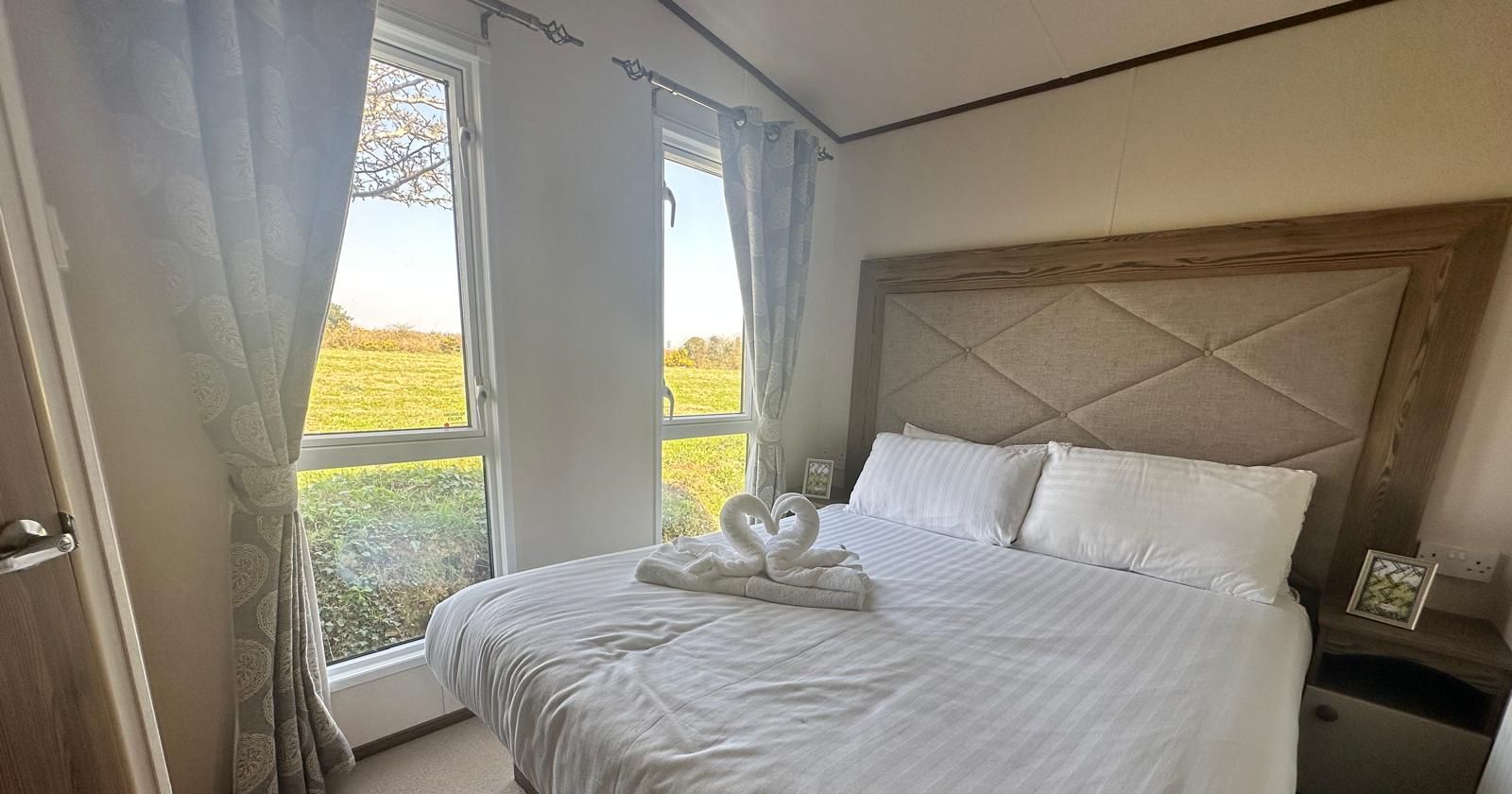 Bright bedroom with a double bed, padded headboard, and towel swans, beside large windows overlooking green fields.