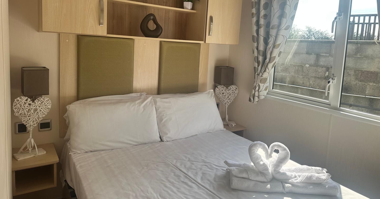 Cosy double bedroom with light wood headboard, built-in shelving, bedside lamps and swan towel display by the window