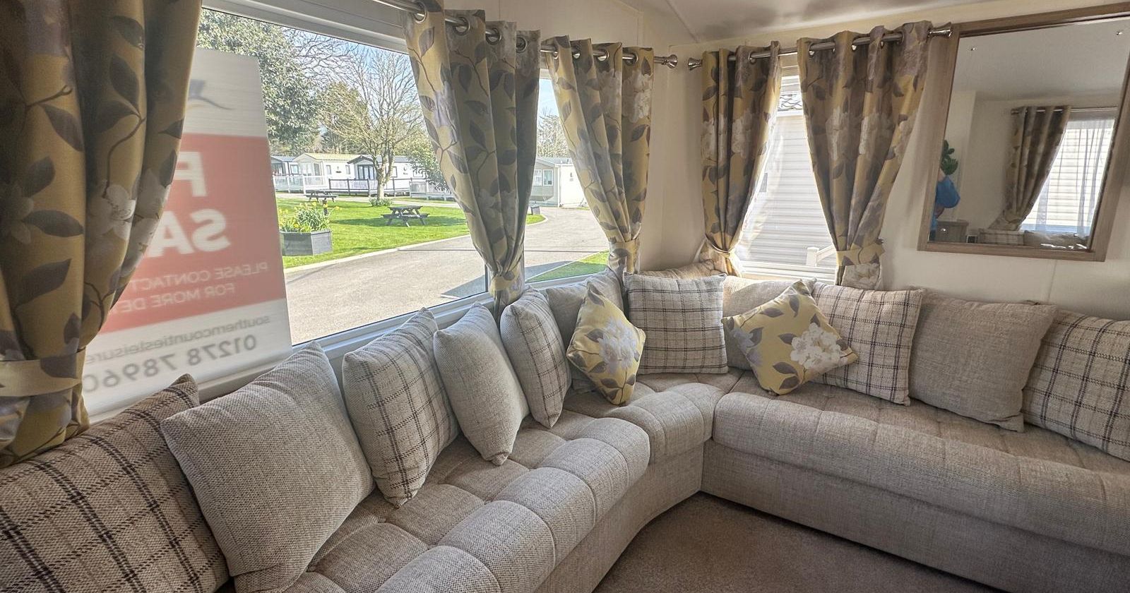 Bright caravan lounge with L-shaped sofa, patterned cushions and floral curtains by large windows