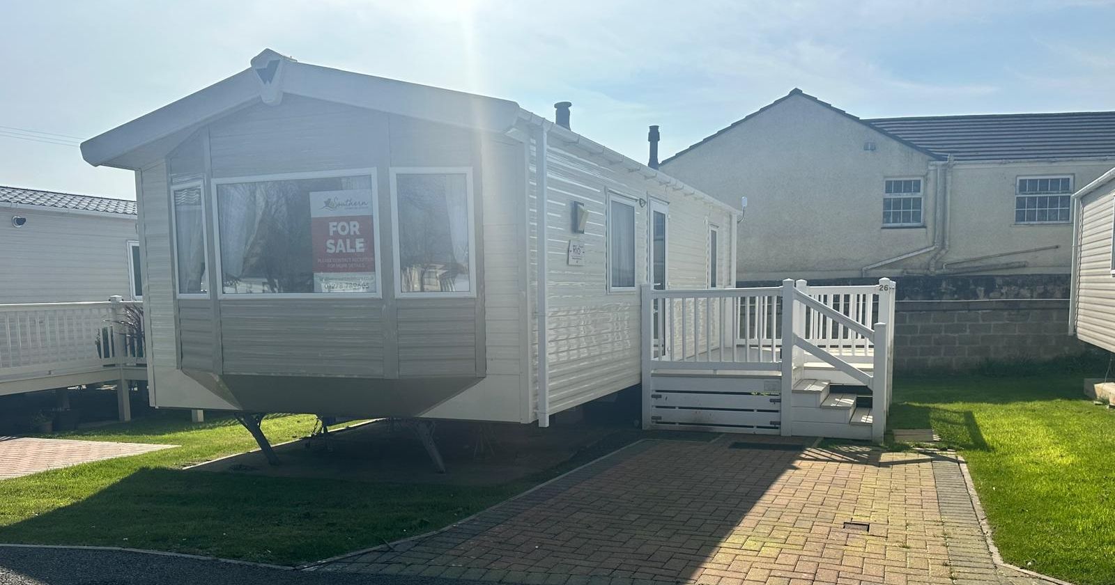 Static caravan with front windows and a white-railed deck, with a for sale sign in the window