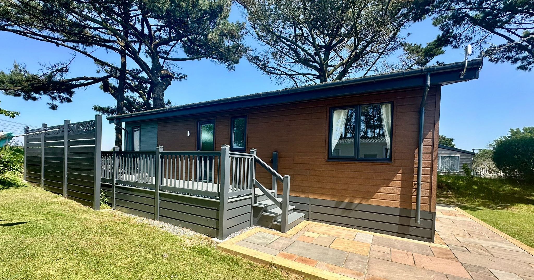 Modern wooden chalet with grey deck and steps in a sunny garden beneath tall pine trees