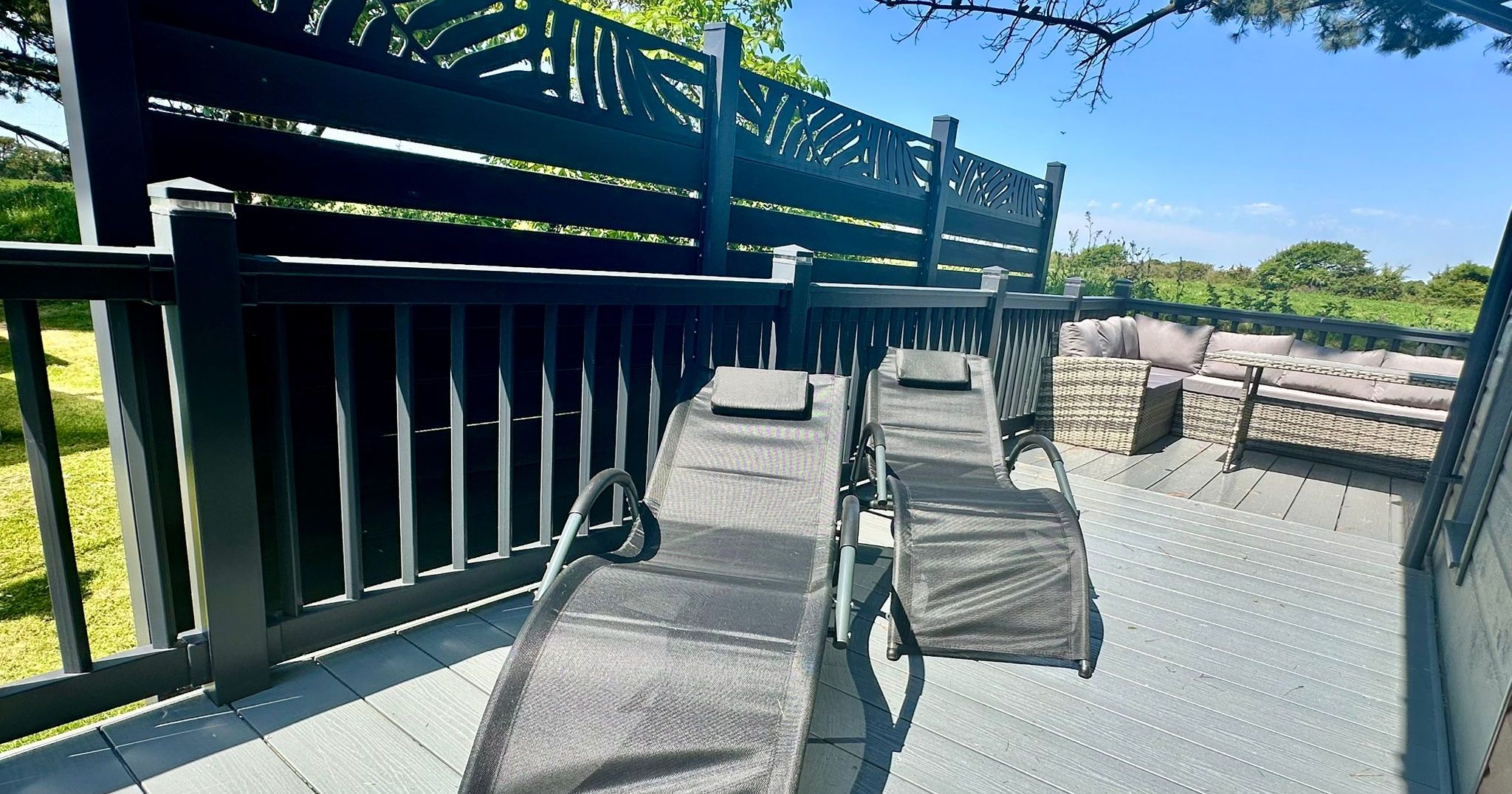 Sunlit deck with two grey sun loungers and a corner rattan sofa set, enclosed by a dark privacy screen and railings.