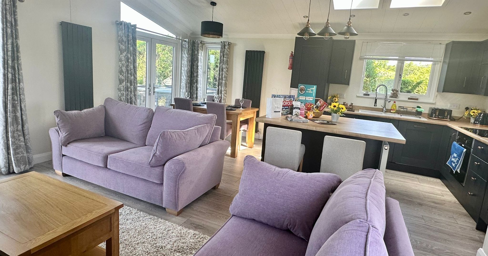 Open-plan living room and kitchen with lilac sofas, wooden dining table and dark grey cabinets under skylights