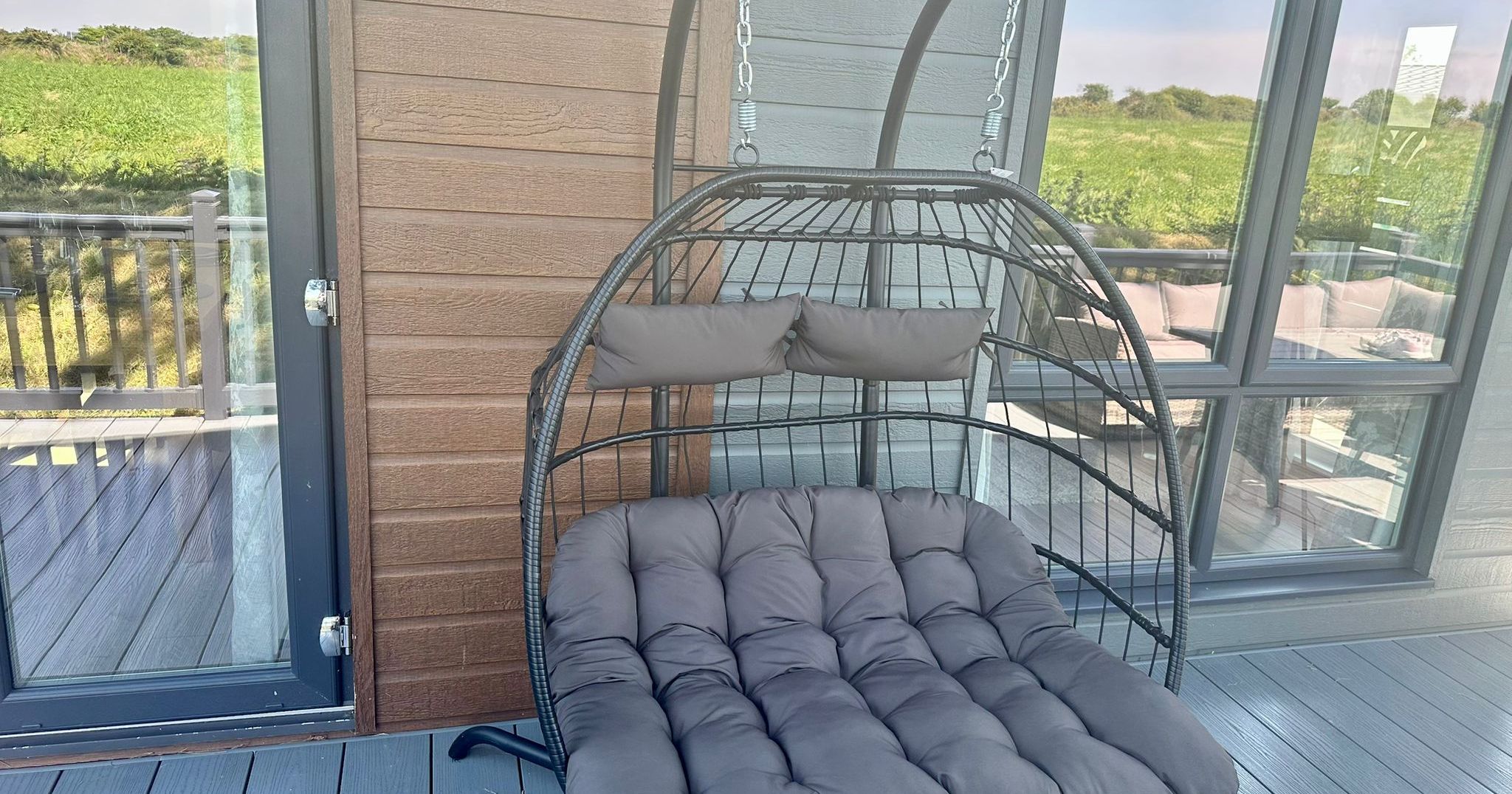 Hanging egg chair with grey cushions on a deck beside a lodge, overlooking green fields.