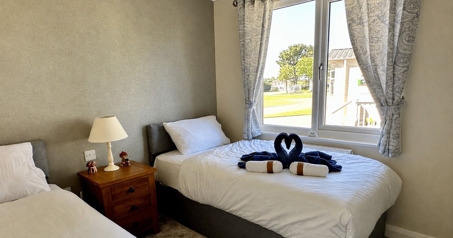 Bright bedroom with two single beds, bedside table and lamp, and a large window with patterned curtains and folded towel swans on the bed.