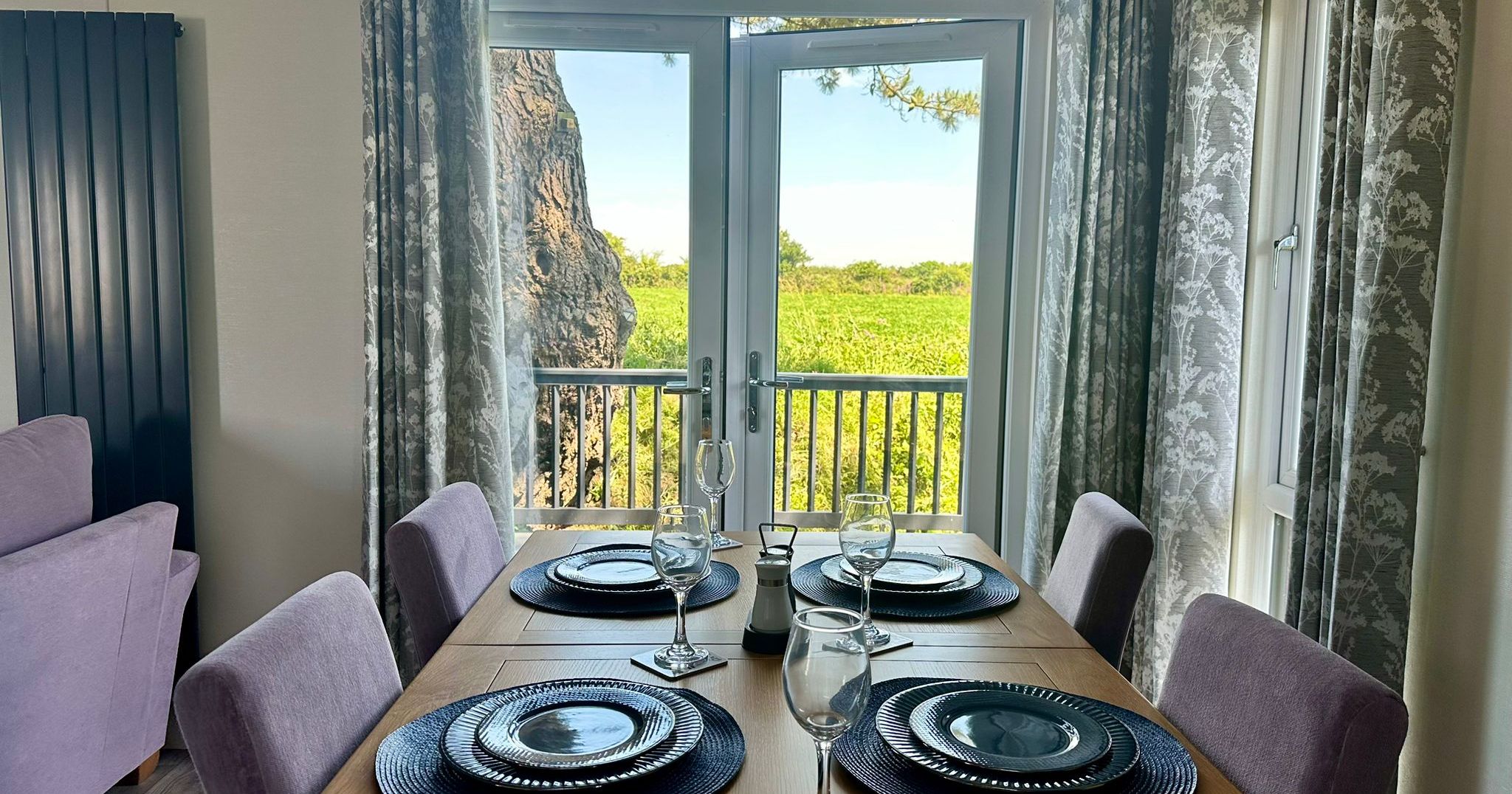 Dining table set with four place settings beside French doors opening onto a balcony with countryside views