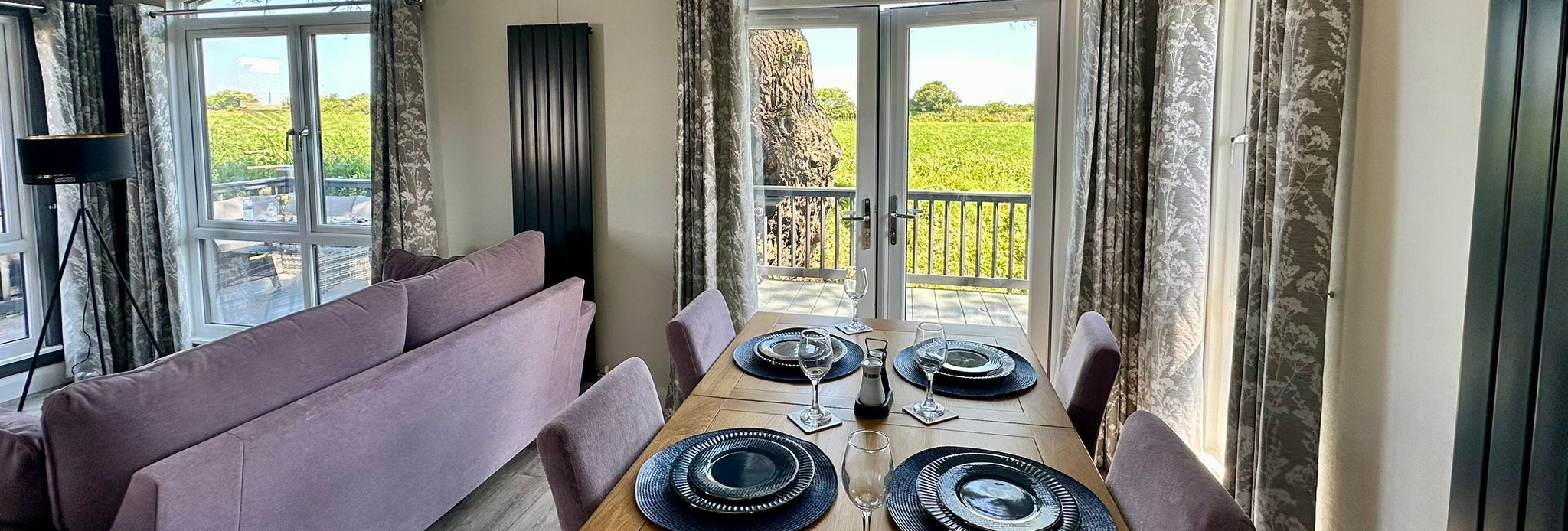 Dining area with a wooden table set for four beside a lilac sofa, with French doors opening to a balcony and countryside views