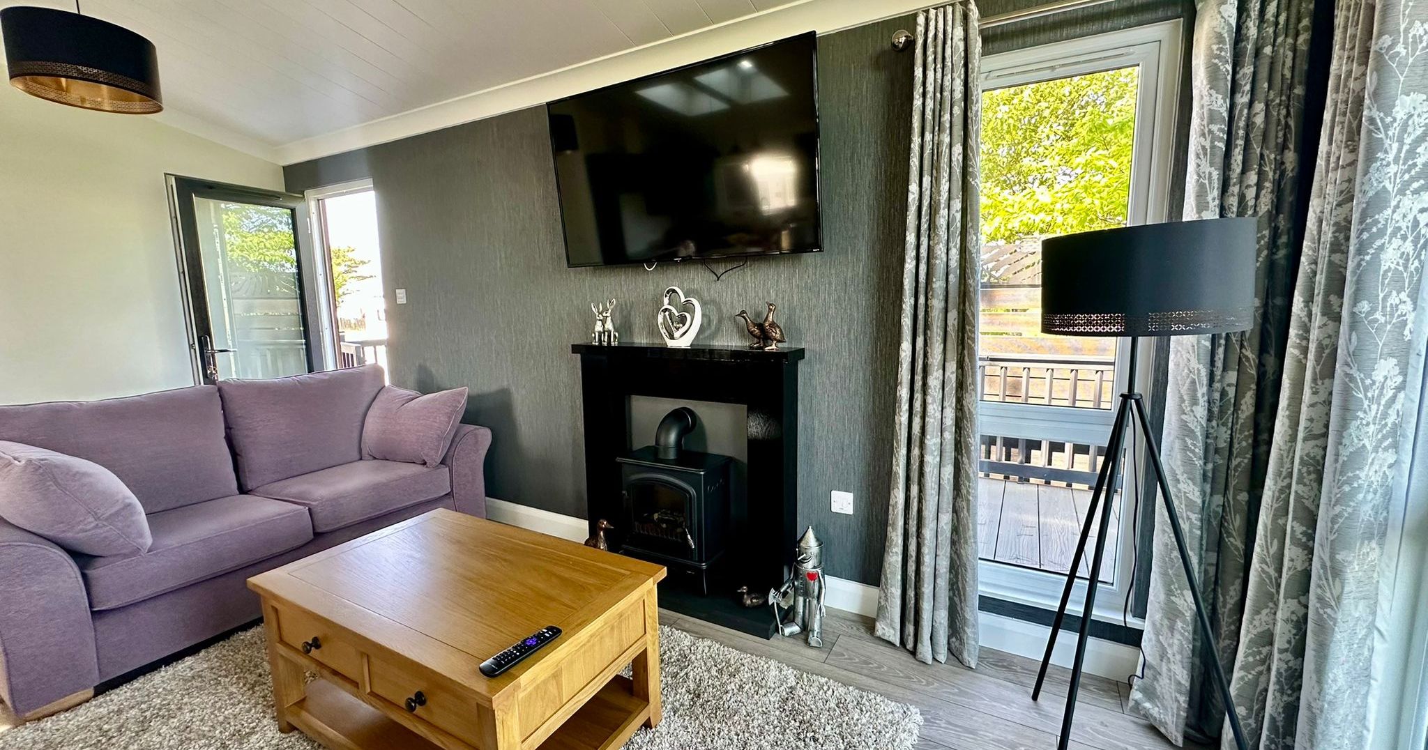 Modern lounge with purple sofa, wooden coffee table, wall-mounted TV above a fireplace, and large windows with grey curtains
