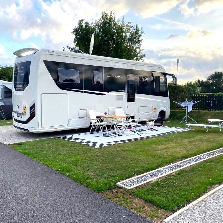White motorhome parked on a campsite pitch with outdoor table, chairs and sun loungers on a chequered mat