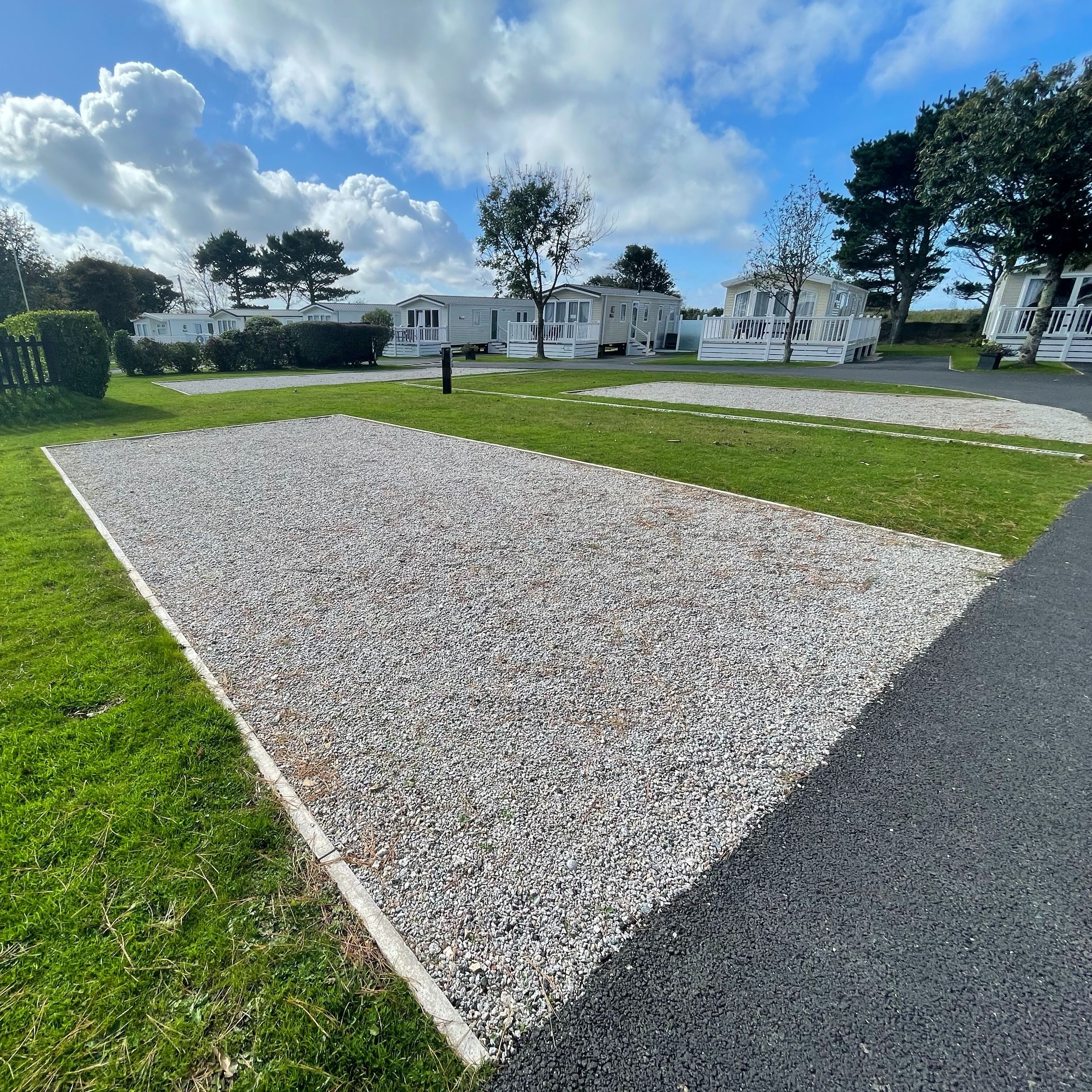 Gravel pitch with electric hook-up on a landscaped holiday park, with static caravans and trees under a blue sky