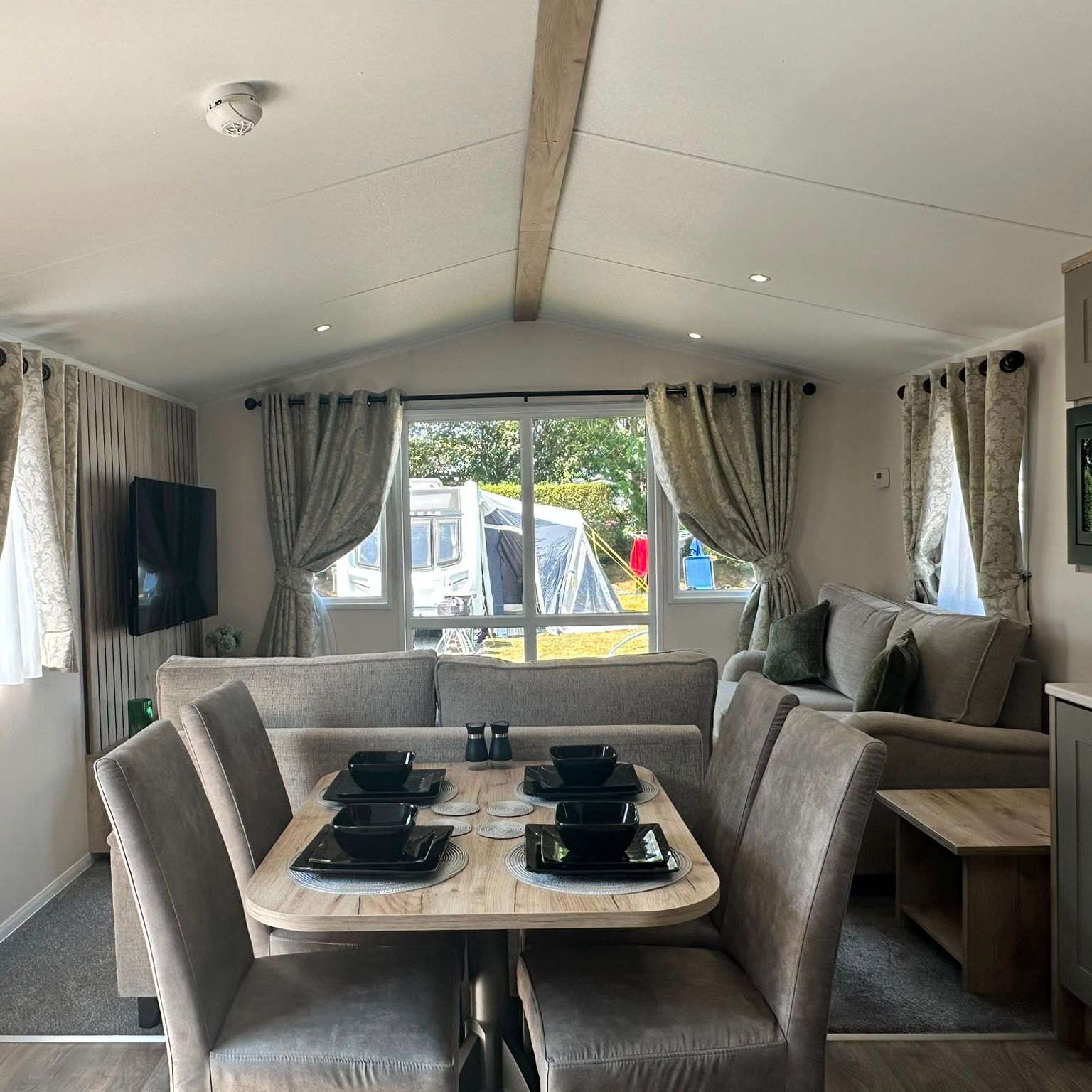 Modern caravan lounge and dining area with grey sofa, dining table set for four, wall-mounted TV, and curtains framing a large window.