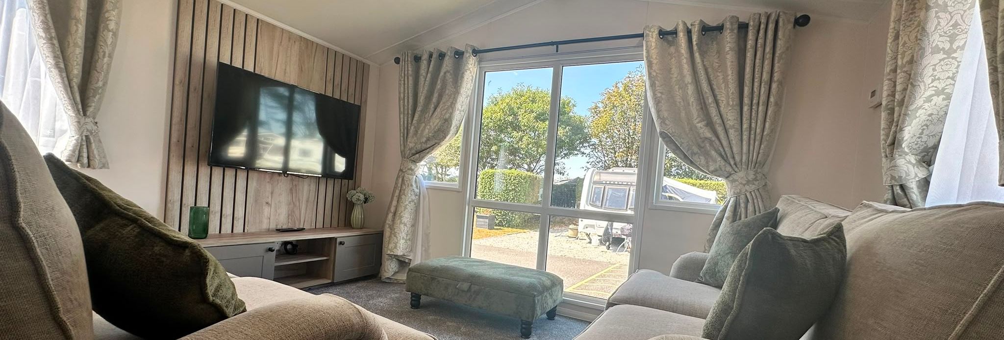 Cosy lounge with two beige sofas, wall-mounted TV and large window with patterned curtains