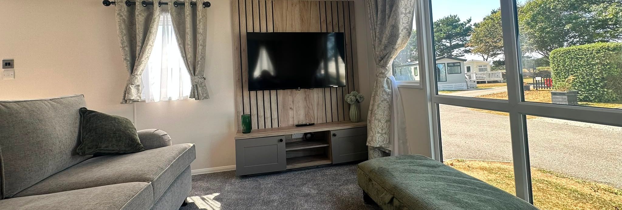 Bright living room with grey sofa, wall-mounted TV and large window with patterned curtains overlooking a holiday park