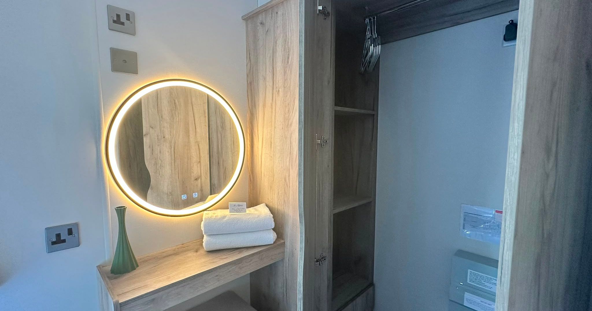 Open wooden wardrobe with hangers beside a vanity with an illuminated round mirror and folded towels