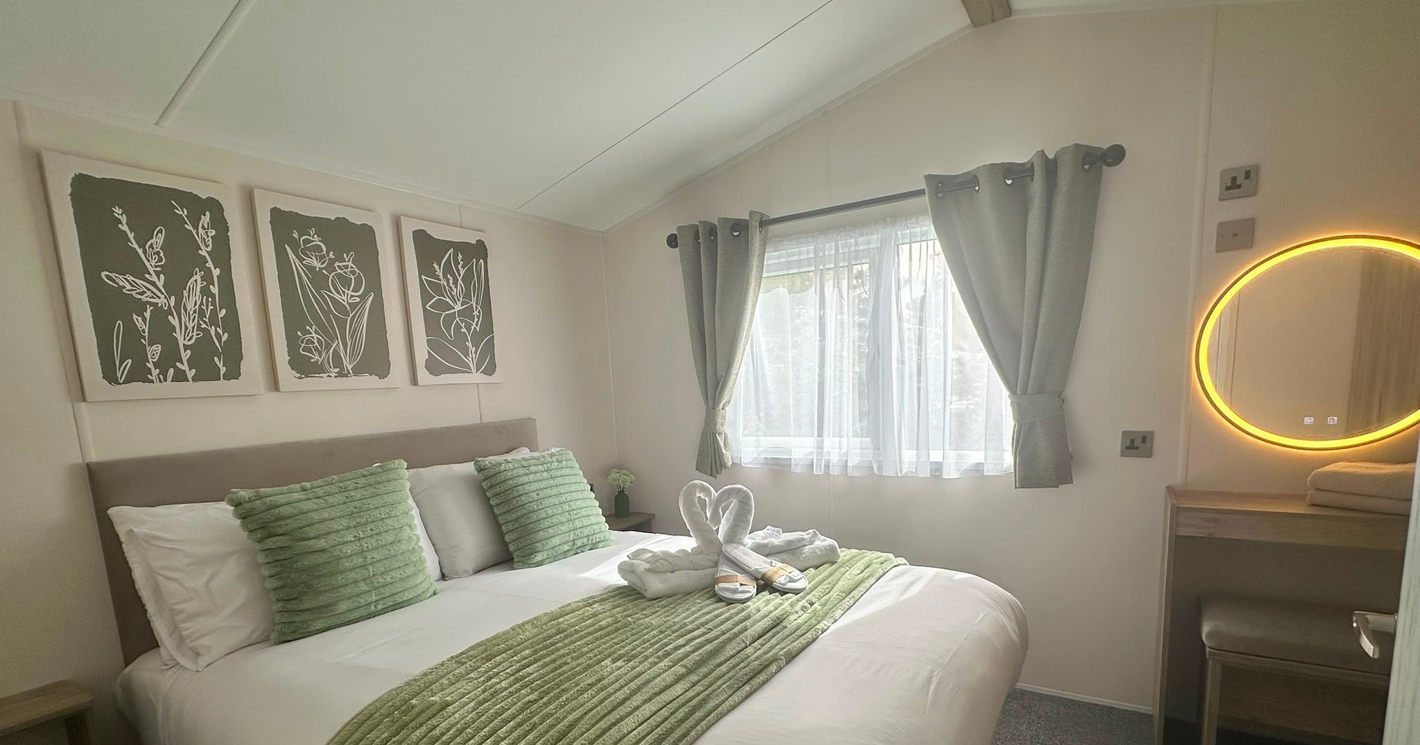 Bright double bedroom with white bedding, green cushions and throw, grey curtains, botanical wall art and illuminated round mirror