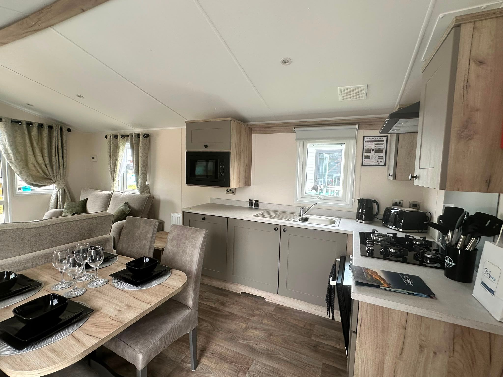 Open-plan caravan kitchen and dining area with grey cabinets, gas hob, sink, microwave, and a table set with plates and glasses