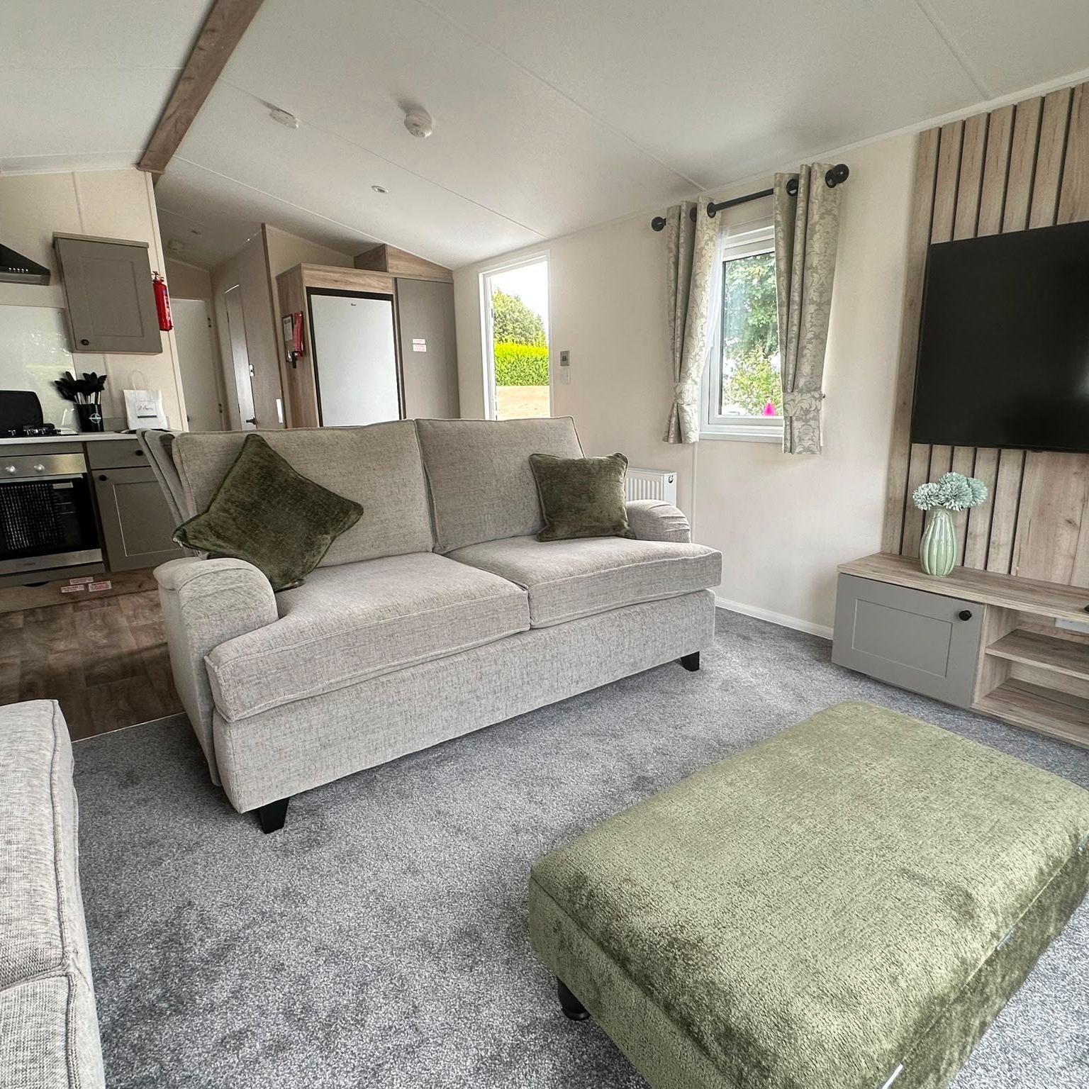 Modern open-plan lounge and kitchen with grey sofas, green footstool and wall-mounted TV