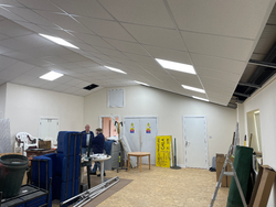 Two people in a room with a partially installed suspended ceiling and various furniture and equipment scattered around.