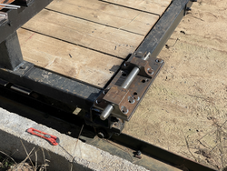 Close-up of a metal hinge mechanism attached to a wooden platform with a red utility knife nearby