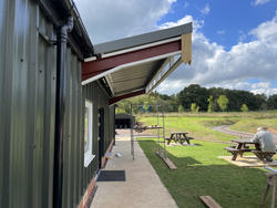 Green metal building with metal awning under construction, scaffolding, picnic benches, grassy area, and a person sitting outside.