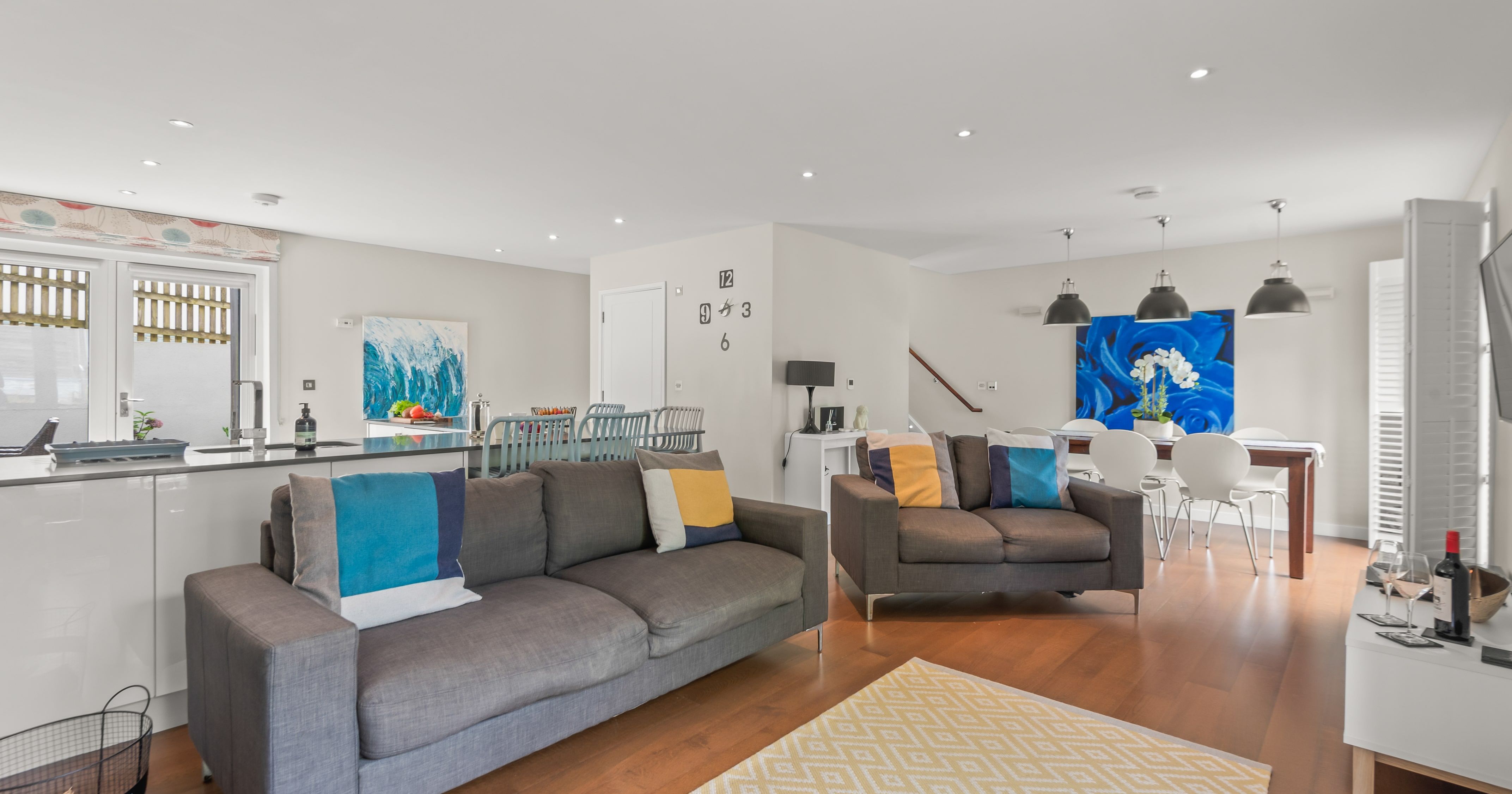 Modern open-plan living and dining area with grey sofas, colorful pillows, wooden floor, white dining table, large blue rose artwork, and contemporary kitchen.