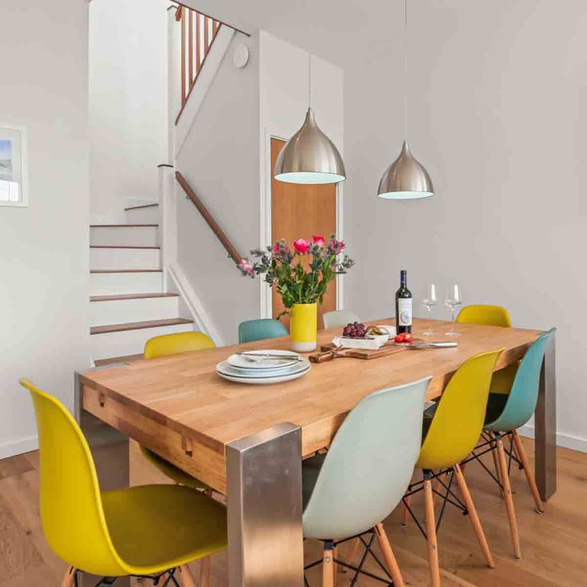 Modern dining room with multicolored chairs, a wooden table, flowers, wine, and staircase in the background