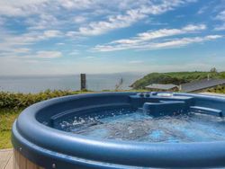 Outdoor hot tub overlooking the ocean and green hills under a blue sky with scattered clouds