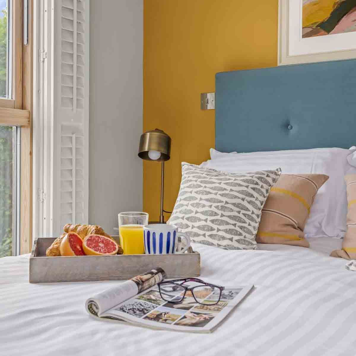 Breakfast tray with croissants, grapefruit, orange juice, and coffee on a bed with patterned pillows and a magazine