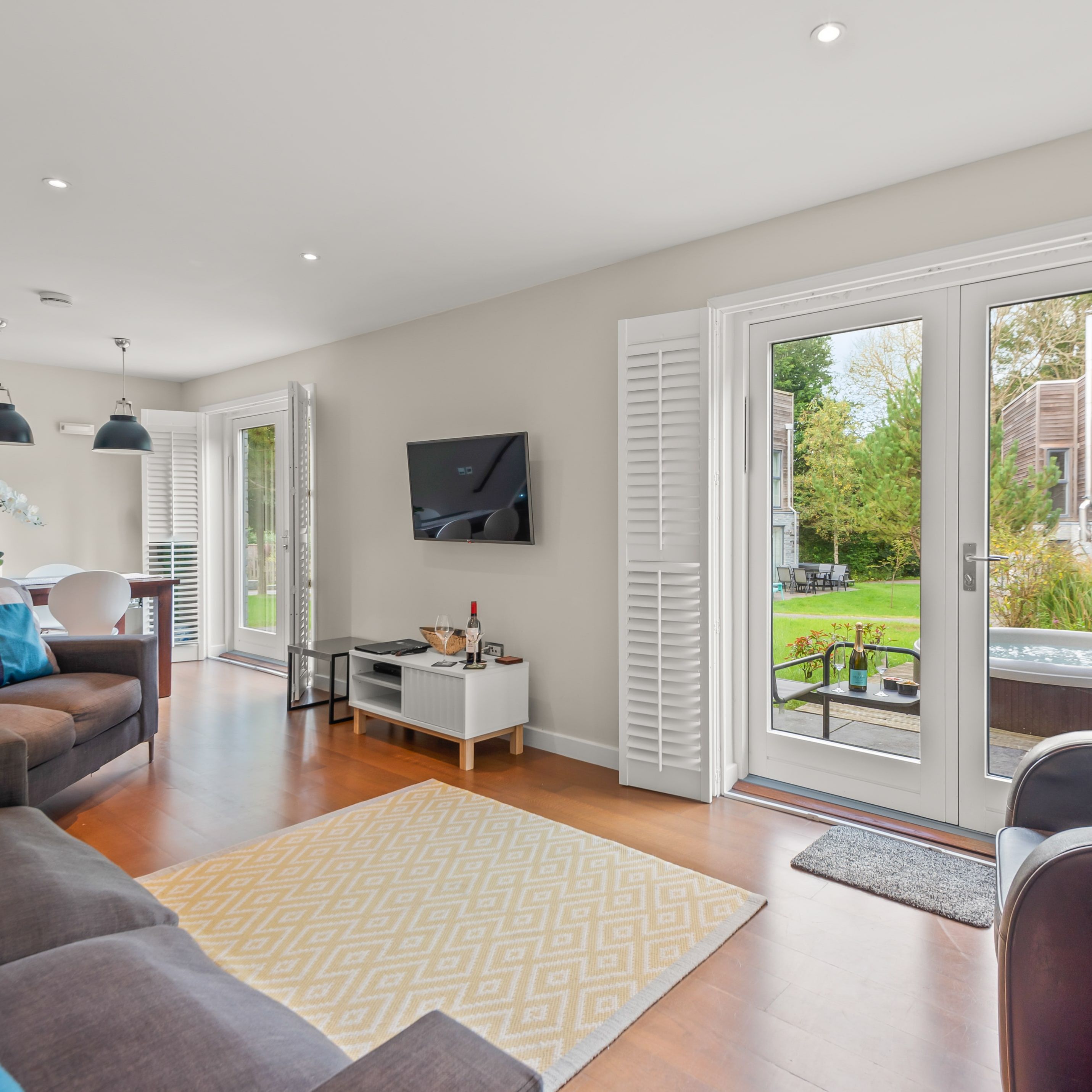 Modern living room with large glass doors opening to a garden with a hot tub, stylish furniture, a TV on the wall, and a dining area with contemporary decor.