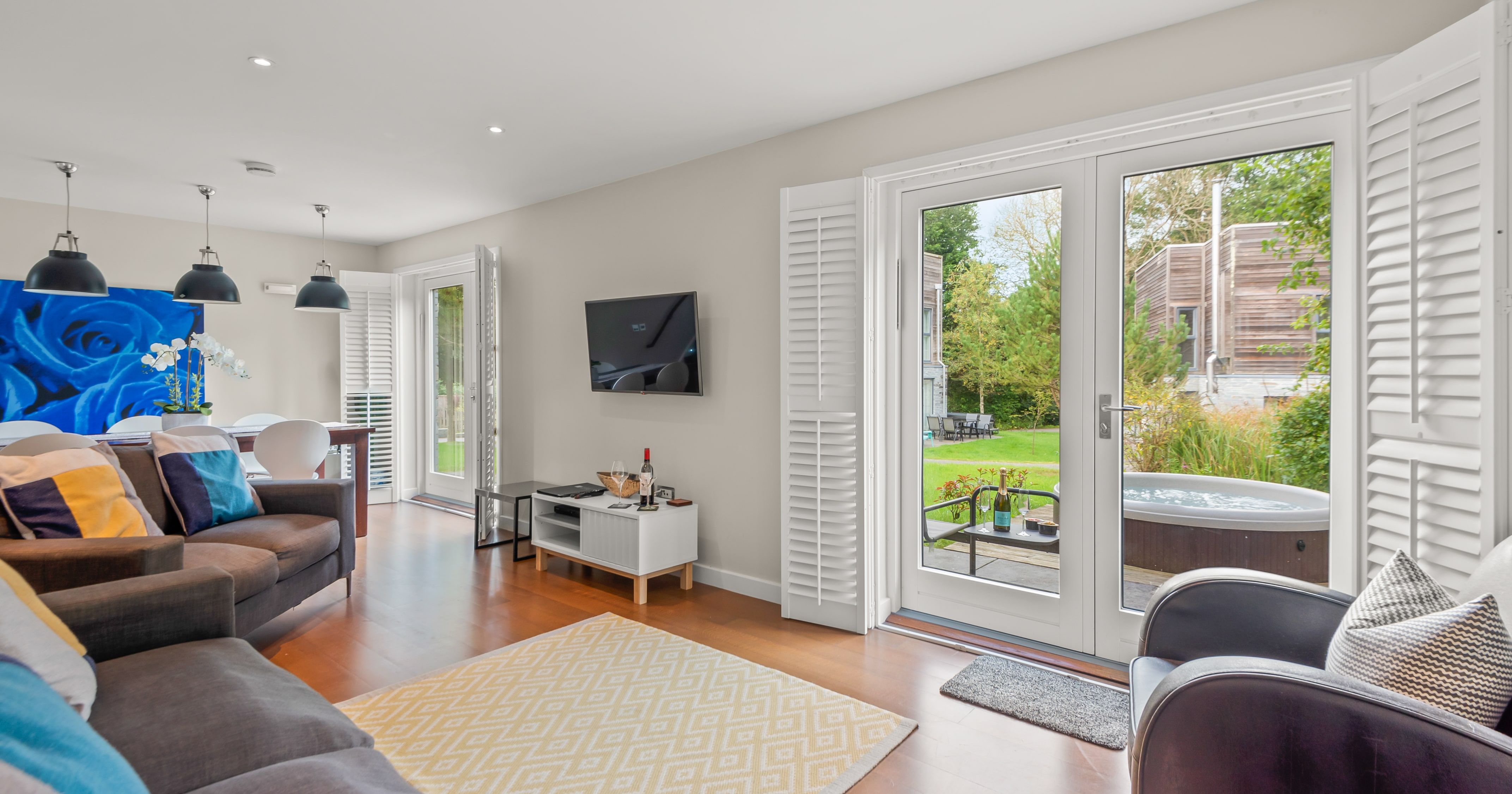 Modern living room with large glass doors opening to a garden with a hot tub, stylish furniture, a TV on the wall, and a dining area with contemporary decor.