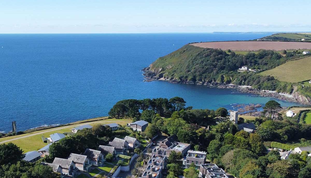 Aerial view of a coastal village with buildings, lush greenery, and cliffs beside a large body of water