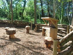 Outdoor woodland play area with log seating and a small wooden playhouse surrounded by a rustic log fence.