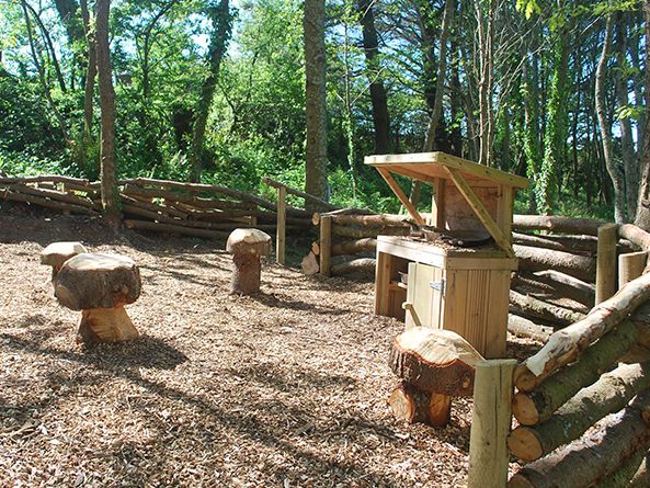Outdoor woodland play area with log seating and a small wooden playhouse surrounded by a rustic log fence.