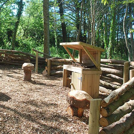 Outdoor woodland play area with log seating and a small wooden playhouse surrounded by a rustic log fence.