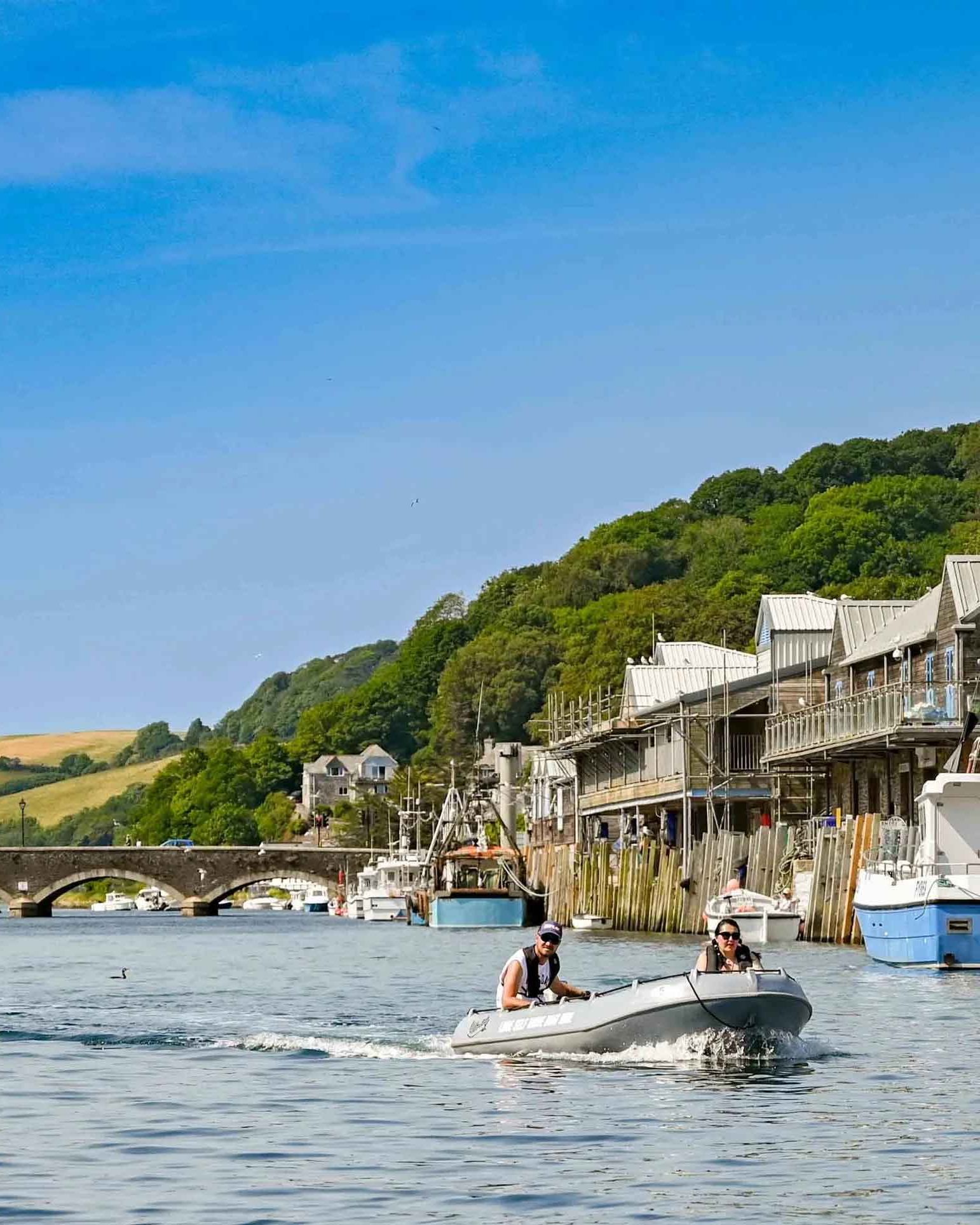 Small boat on a river with people, surrounded by buildings, docks, and moored boats, with a stone bridge and green hills in the background.