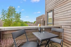 Modern balcony with two chairs and a table set for drinks overlooking greenery and contemporary wooden buildings.