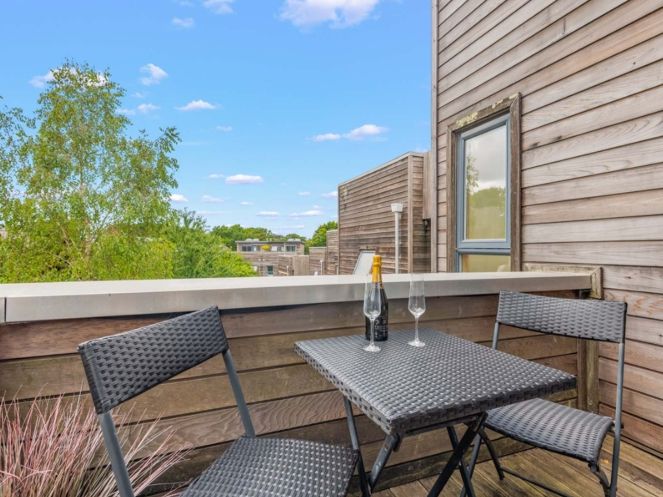 Modern balcony with two chairs and a table set for drinks overlooking greenery and contemporary wooden buildings.