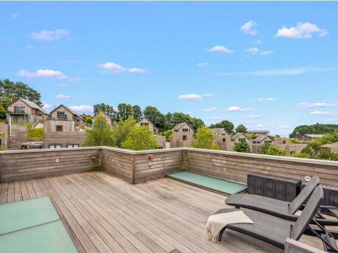 Modern rooftop terrace with wooden decking, lounge chairs, and views of contemporary houses surrounded by greenery.