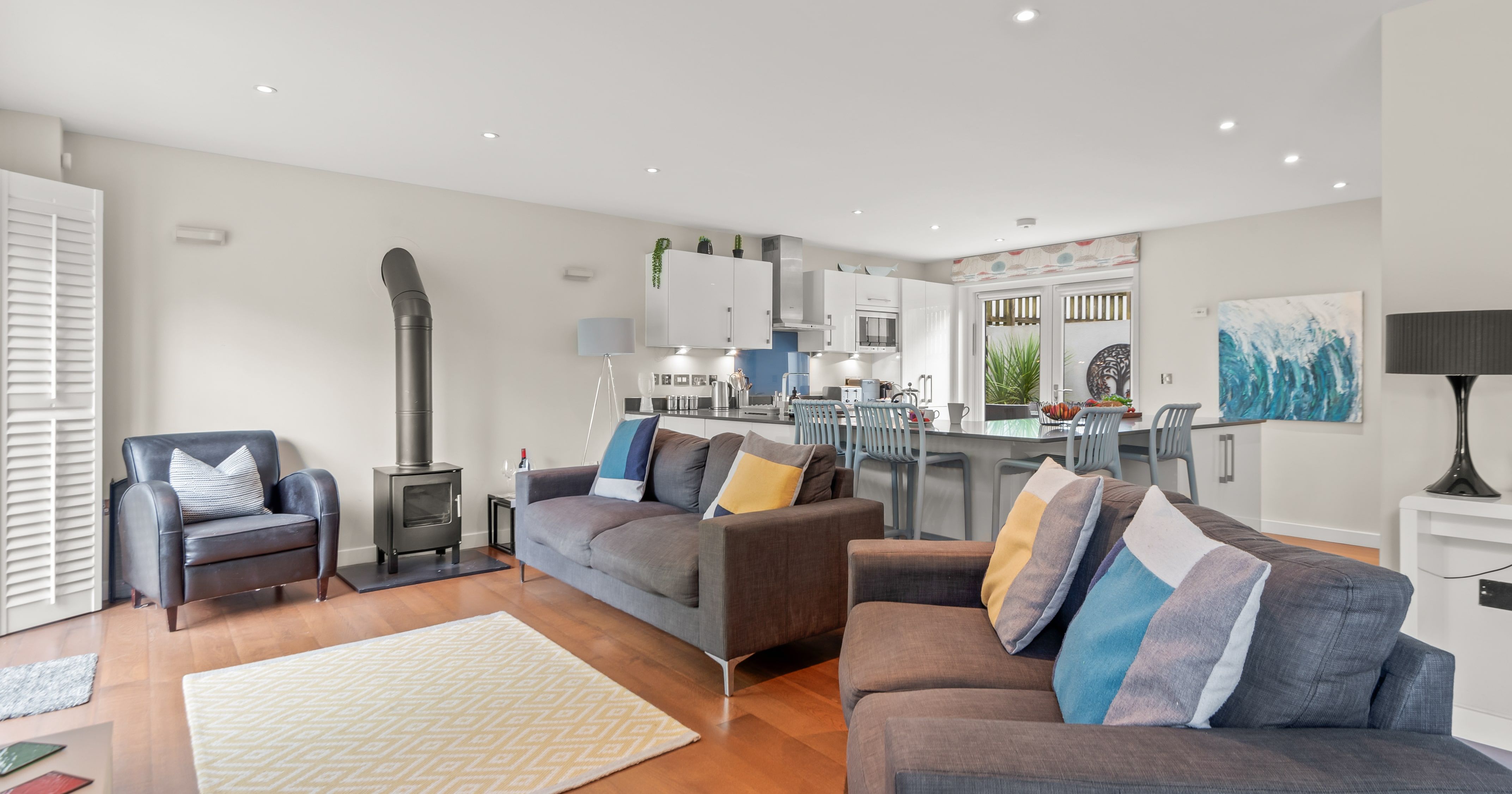 Modern open-plan living room and kitchen with grey sofas, colorful cushions, wood-burning stove, armchair, white cabinets, breakfast bar with blue chairs, and contemporary decor.