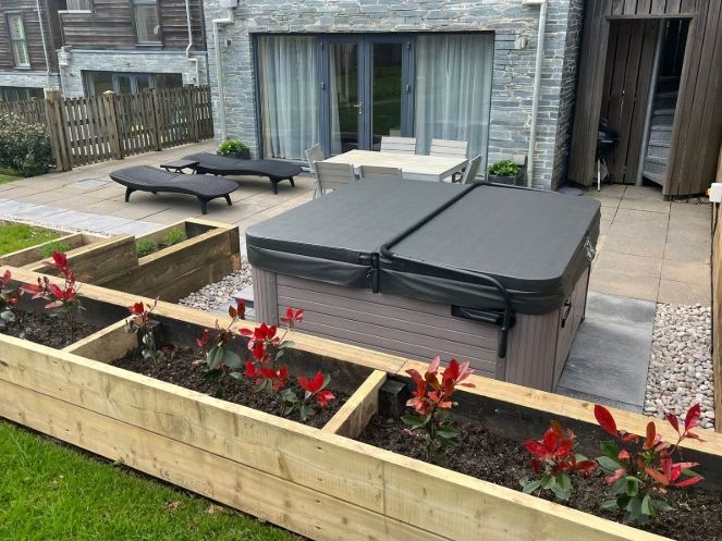 Outdoor patio area with a closed hot tub, sun loungers, table and chairs, and raised wooden garden beds with red-leafed plants.