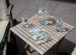 Wooden table with a glass jug of water, two glasses of water, and a handbook outdoors