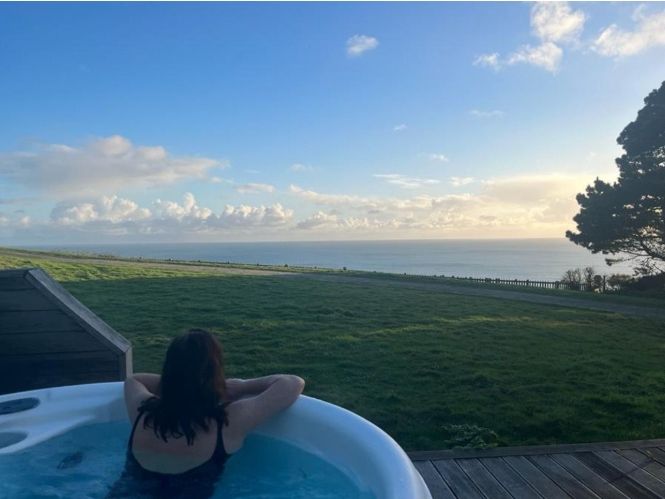 Person relaxing in a hot tub overlooking a grassy field with ocean and sky in the background.