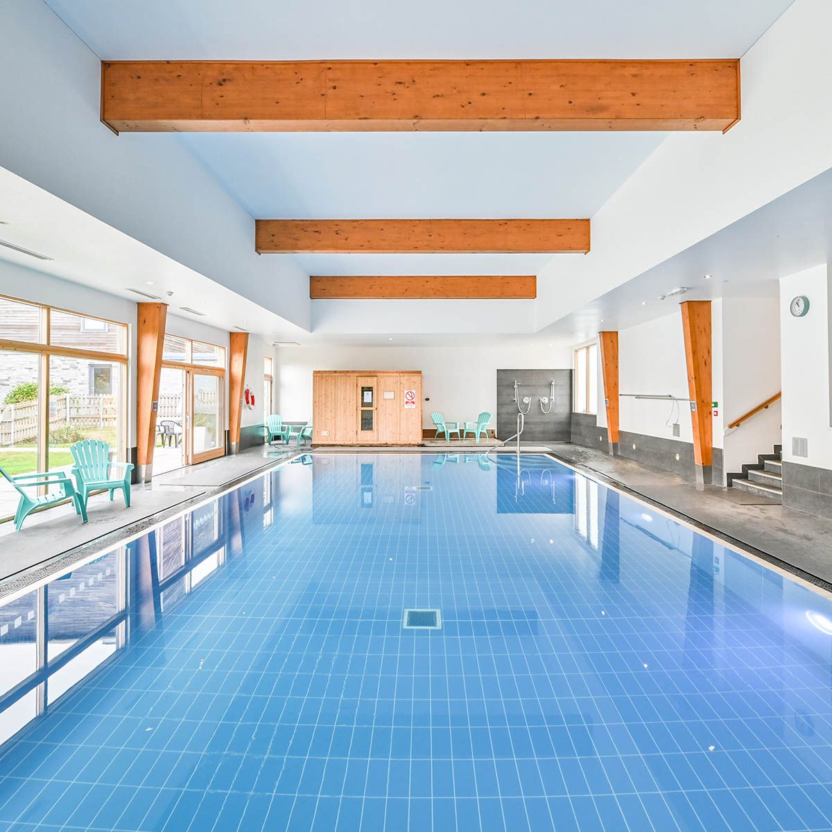 Modern indoor swimming pool with wooden beams, large windows, and poolside chairs