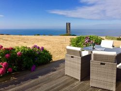Outdoor dining area on a wooden deck overlooking the ocean, with flowers and a field in the background.