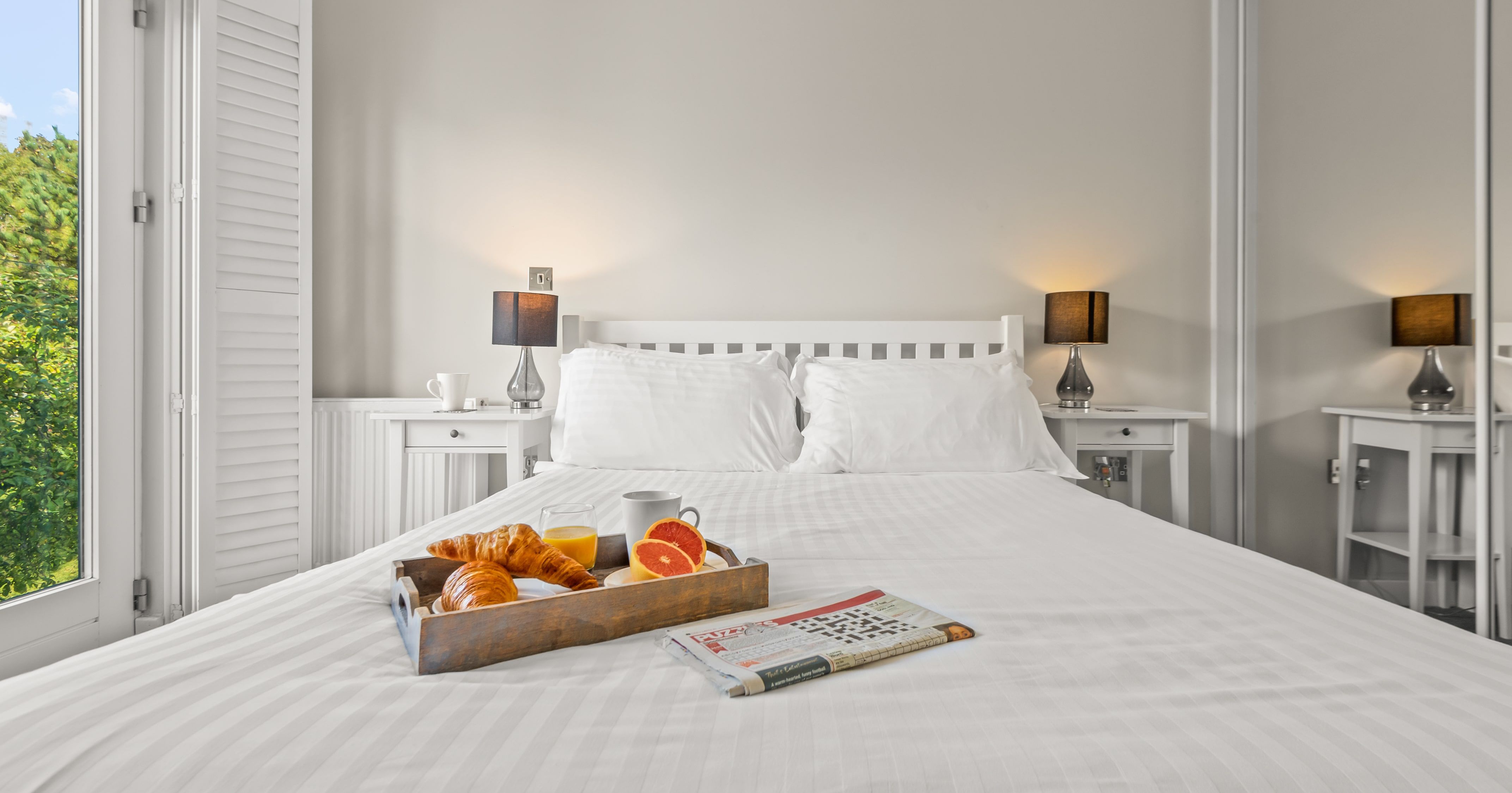 Bright bedroom with white bedding, bedside tables with lamps, a large window, and a breakfast tray with croissants, orange juice, coffee, and fruit on the bed with a newspaper.