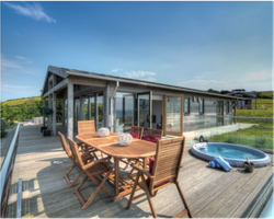 Modern wooden house with large glass windows, an outdoor dining table, and a hot tub on a spacious deck overlooking green hills.